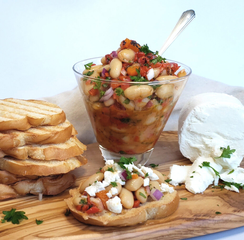 A rustic wooden charcuterie board features the following from left to right: a stack of toasted Italian bread, a small glass bowl of roasted red pepper and cannellini bruschetta and a creamy slice of fresh goat cheese. In the forefront of the picture rests a piece of crusty bread piled high with bruschetta and topped with goat cheese crumbles.