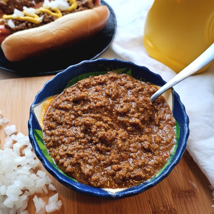 A bowl of Easy Detroit Style Coney Dog Sauce sits amidst a pile of diced sweet onions, a bottle of mustard and two perfectly topped hot dogs smothered in Coney Sauce, onions and mustard.