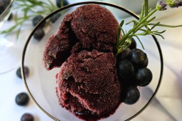 A scoop of this Heavenly Lavender French 75 Blueberry Sorbet is topped with fresh blueberries and lavender flowers.