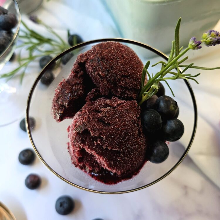 A scoop of this Heavenly Lavender French 75 Blueberry Sorbet is topped with fresh blueberries and lavender flowers.