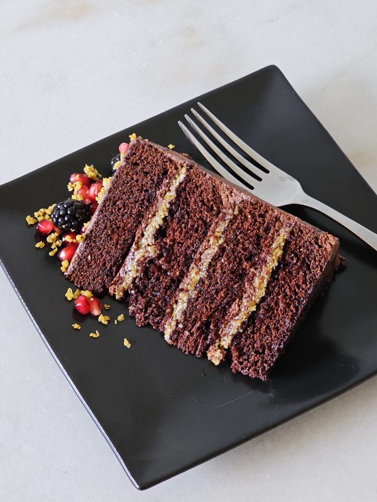A slice of Dubai Chocolate Cake reveals alternating layers of chocolate cake brushed in a chocolate simple syrup, Dubai chocolate filling, and whipped chocolate Ganache. The cake is finished in whipped Ganache and adorned with fresh fruit.