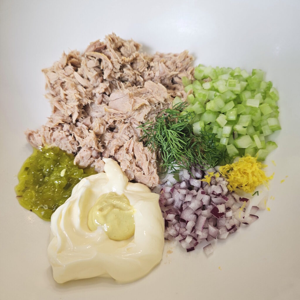 A bowl containing all the ingredients in this homemade tuna salad recipe. Tuna, fresh celery, red onion, lemon zest, sweet pickle relish, mayonnaise, Dijon mustard, salt and pepper. 