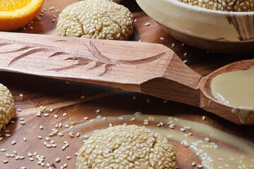 A bowl of freshly baked Easy Orange Cardamom Tahini Cookies share a platter with a fresh orange, sesame seeds, and Tahini.