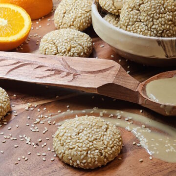 A bowl of freshly baked Easy Orange Cardamom Tahini Cookies share a platter with a fresh orange, sesame seeds, and Tahini.