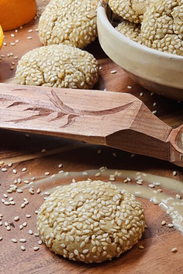 A bowl of freshly baked Easy Orange Cardamom Tahini Cookies share a platter with a fresh orange, sesame seeds, and Tahini.